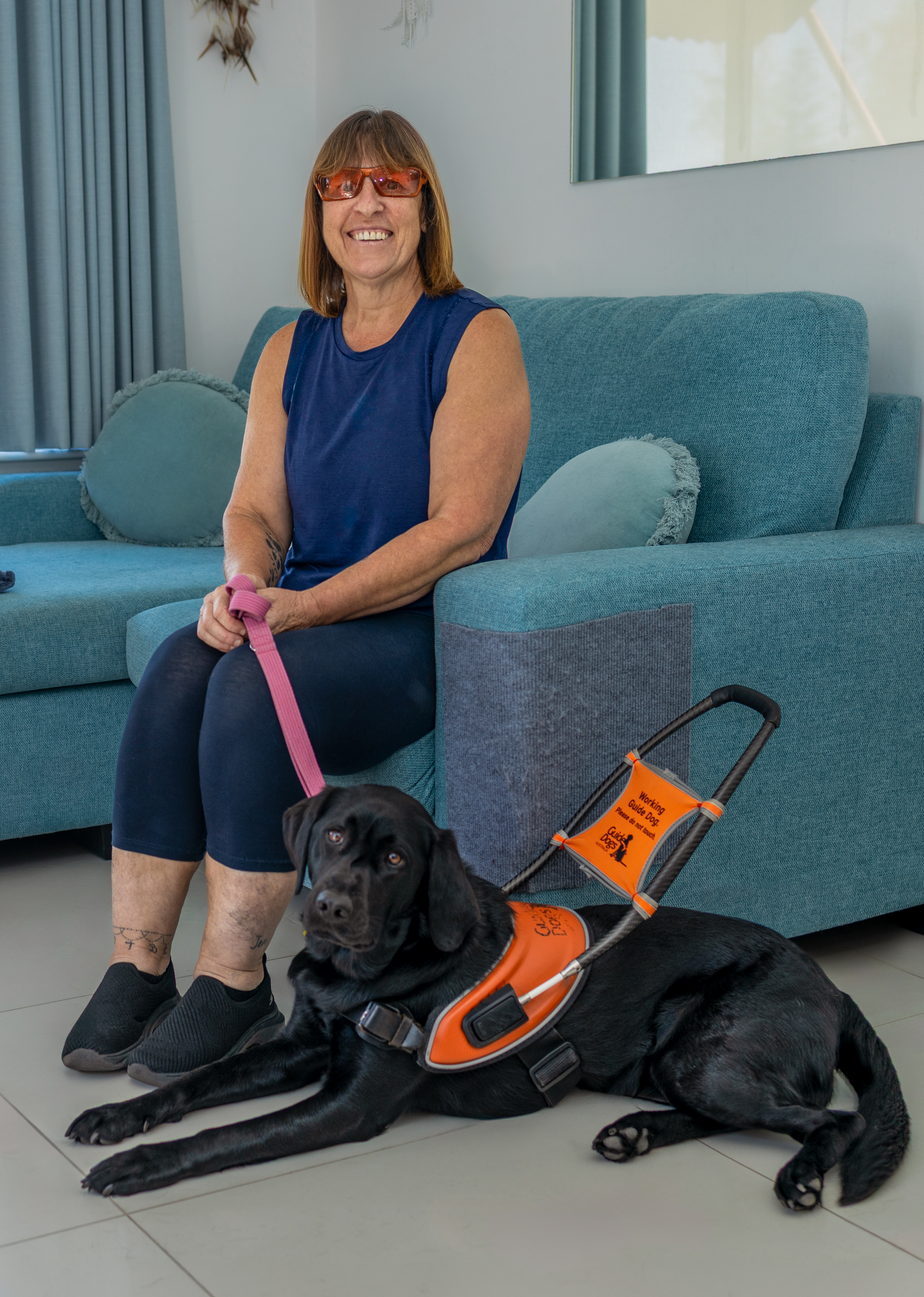 Image shows Penny sitting on a chair next to a black Guide Dog in an orange harness.